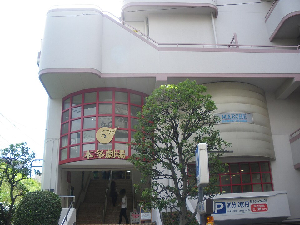 白い建物の赤い窓に本多劇場の看板があり、道路から入口の階段が見える外観
