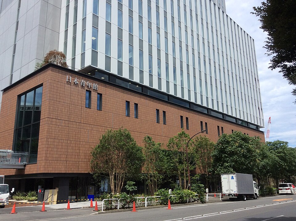 日本青年館の看板がある赤茶色の低層部と、その上に続く銀色の外壁を通り側から見た現建物