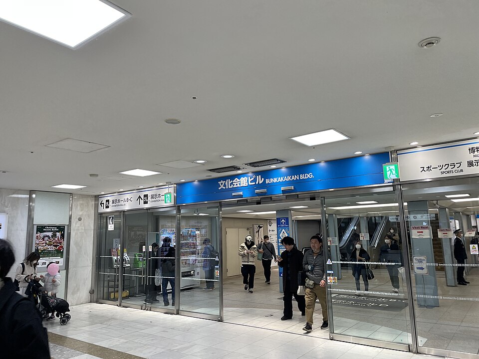 文化会館ビルと書かれた青い案内看板とガラスの入口、その先のエスカレーター