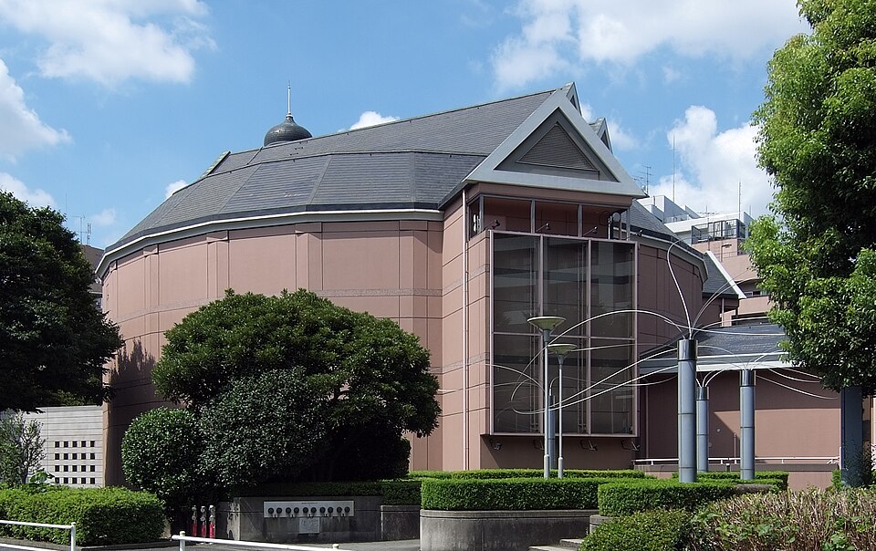 円形に広がる東京グローブ座の劇場と三角形の屋根、ガラス面や周囲の木々が見える外観